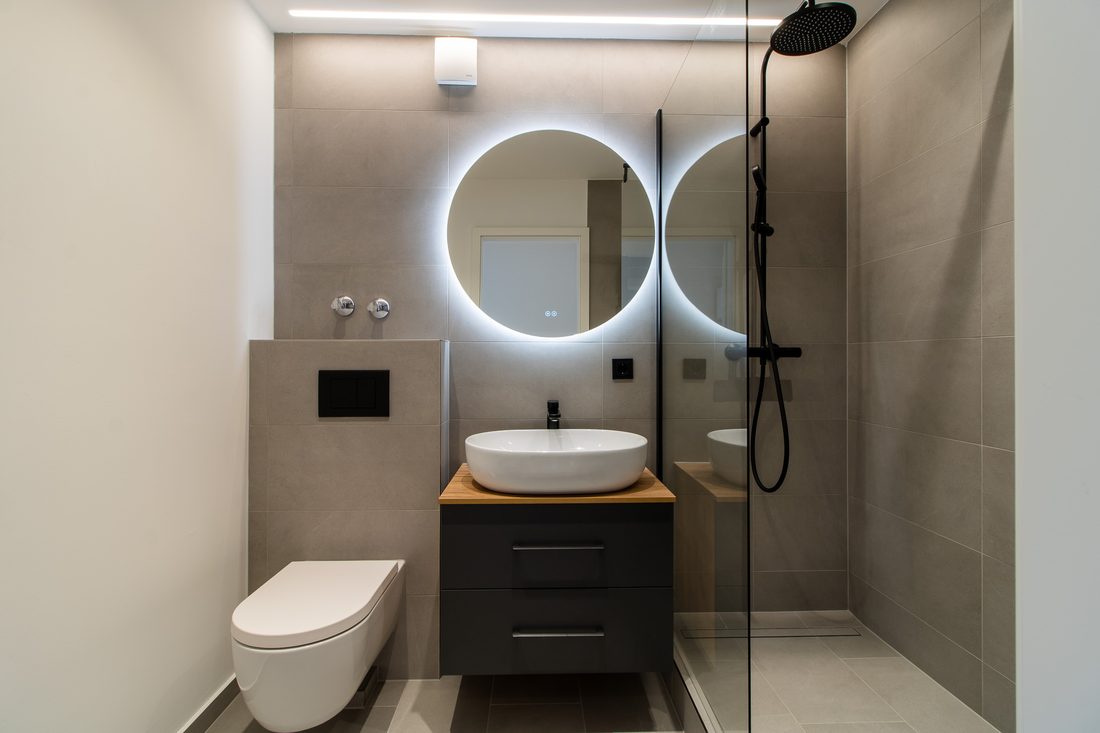 Dasselbe Bad nach der Sanierung: Wand-WC, Aufsatzwaschbecken auf Eichenplatte, runder Spiegel mit Hinterleuchtung und bodengleiche Dusche hinter Glas.