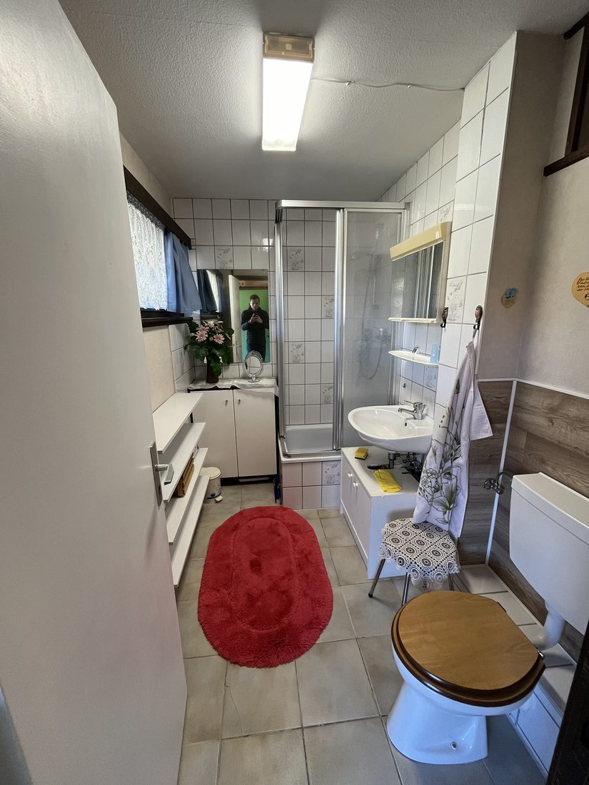 Bad der Ferienwohnung im Chiemgauer Voralpenland vor der Sanierung: weiße Fliesen, enge Duschkabine, alte Sanitärobjekte.